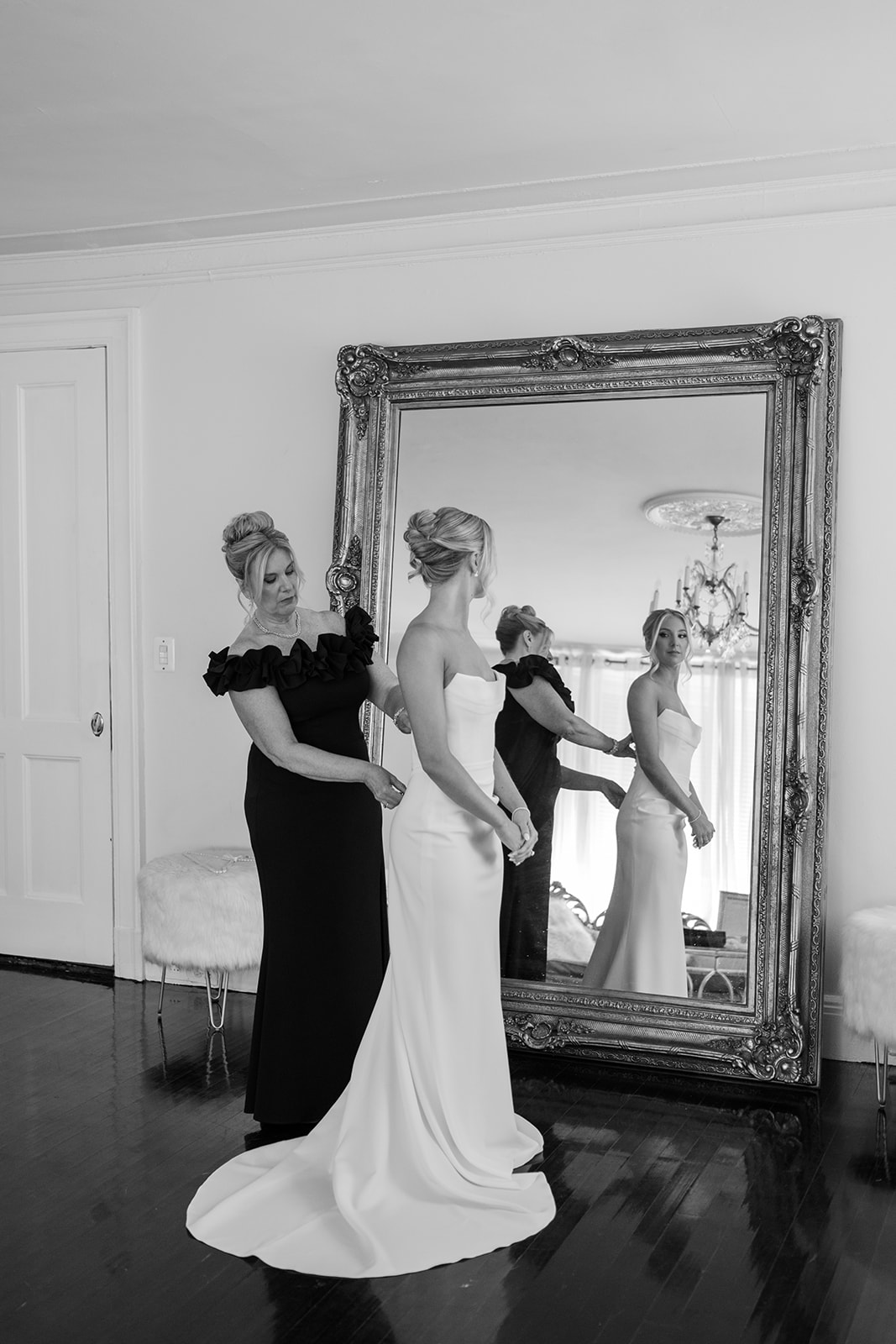 Black and white photo of the mother of the bride helping button the bride's gown in front of a mirror.